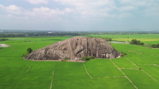 Drone pull-away shot from a towering inselberg in Phu Dien, revealing vast green rice fields across the rural landscape of Southern Vietnam. Hidden gem of natural landmark.