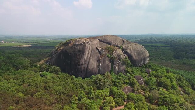 Drone pull-away revealing a massive inselberg rising from dense forest in Phu Dien, Southern Vietnam. Scenic aerial view of an isolated rock hill in tropical countryside.