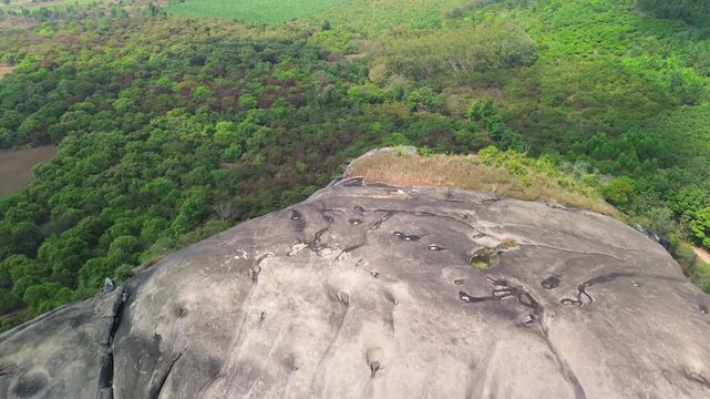 Drone pulling away from the summit of a massive inselberg to reveal the full rock formation rising above dense forest in Phu Dien, Southern Vietnam.