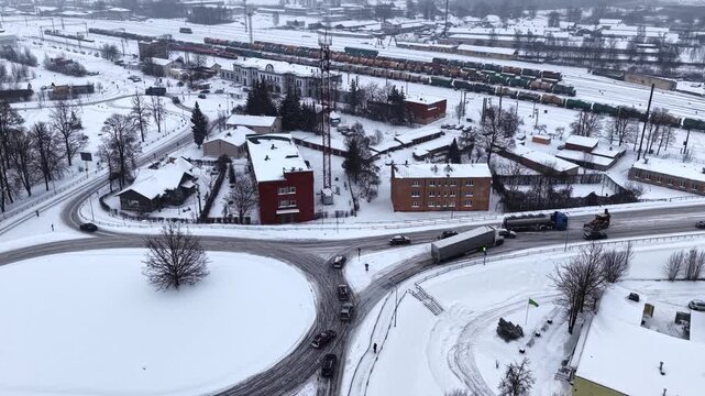 Drone view of a jackknifed semi trailer on an icy curved intersection in a snowy city, with traffic passing and recovery work near a railway yard.