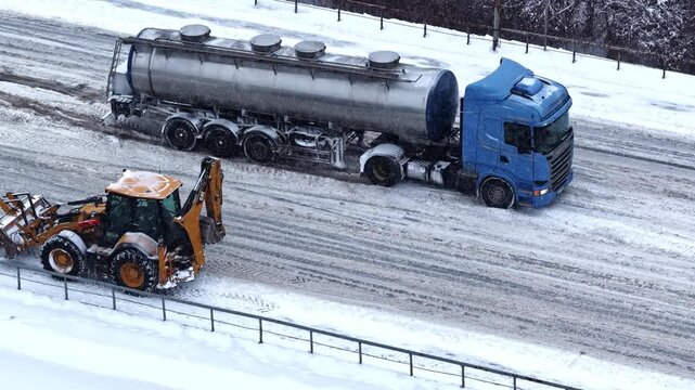 Drone aerial of a blue fuel tanker stuck on an icy winter roadway while a snowplow backhoe works nearby, highlighting hazardous traffic conditions and road maintenance.