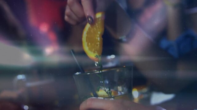 Female patron hands picking herb and lowering orange wedge into cocktail, stirring garnish on bar