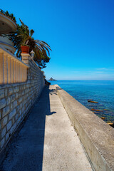 Fototapeta premium Sunlit promenade along the blue Adriatic sea coast in Petrovac, Montenegro with a view of a distant rocky island.