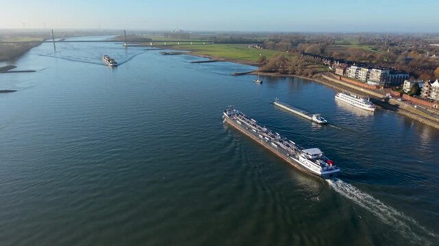 Aerial view of cargo barges and a tanker ship navigating a wide river in Europe. A modern cable-stayed bridge, riverside buildings, and green embankments under a clear blue sky on a sunny day.