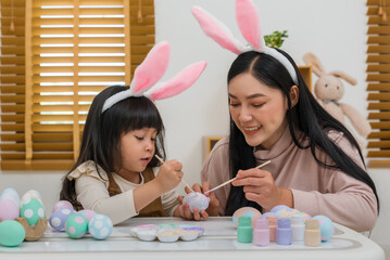 mother and preschool child girl wearing bunny ears painting and decorating egg  together at home on...