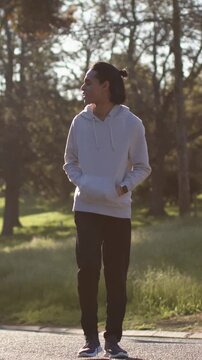 Vertical video: Walking man stopping, scanning trees on park path, sunlight drawing gaze, hoodie