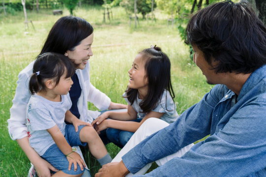 A family enjoys a sunny day outdoors, with parents and two young daughters sharing laughter and conversation while sitting on the grass in a park.