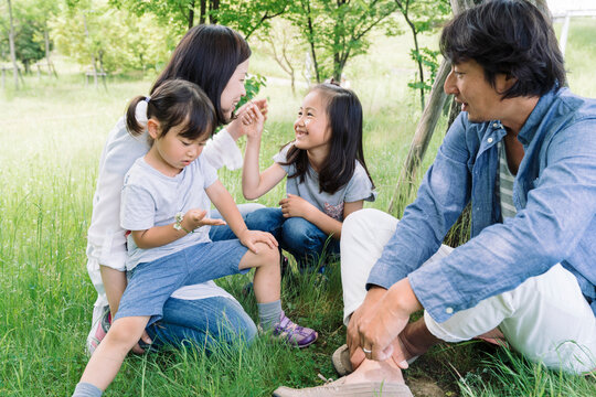 A family enjoys a moment together outdoors, with parents interacting with their two young daughters in a grassy, tree-lined area.