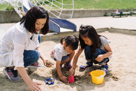 A mother and her two daughters play in a sandbox, building with sand and toys.