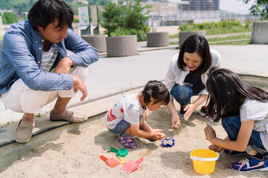 A family enjoys playing in a sandbox together, with parents supervising their children as they build with sand and toys.