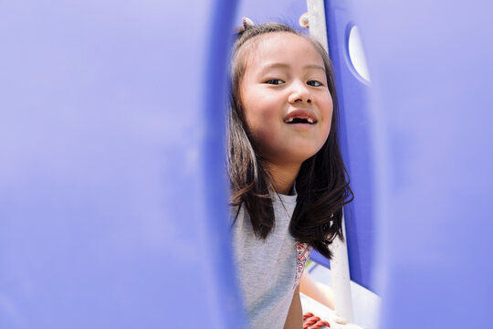 A young girl with missing teeth peeks through a purple tunnel on a playground, her face lit by the sun.