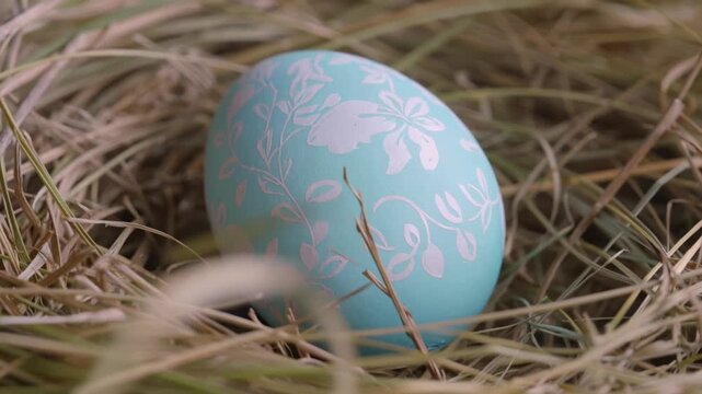 Single pastel blue Easter egg with delicate white floral pattern resting in dry grass nest in macro close-up
