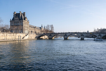 Obraz premium Scenic view of the louvre museum and pont royal bridge over the seine river in paris featuring classical architecture under a clear blue sky during a sunny winter morning with a tourist boat