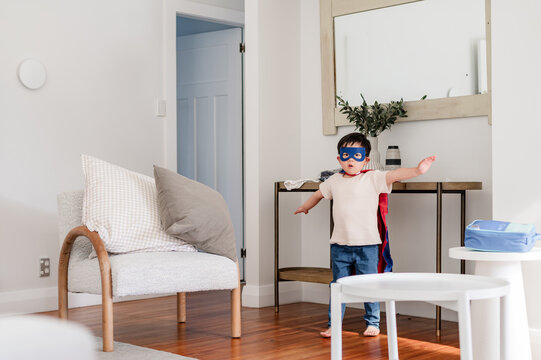 A child with a mask and cape stands in a living room pretending to be a superhero
