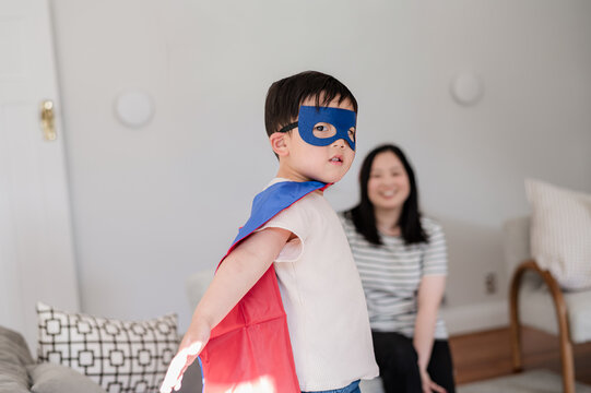 Young boy dressed as a superhero stands proudly in the living room while mother smiles at him.