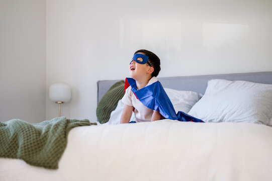 Asian boy wearing superhero costume sitting on the bed in bright bedroom
