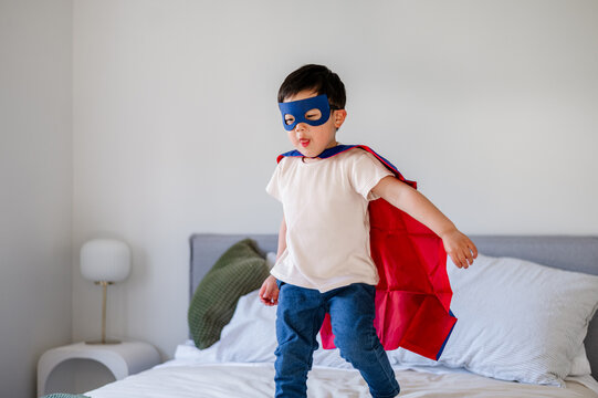 A boy wearing a mask and cape stands on a bed while pretending to be a superhero
