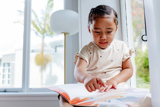 Young samoan girl reading a book while sitting by the windowsill