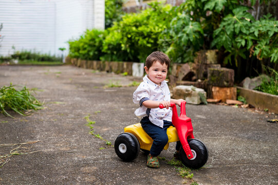Child enjoys riding a red and yellow toy tricycle in a garden area while smiling
