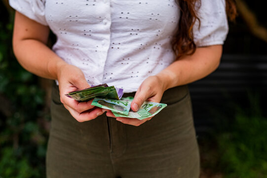 A woman stands outdoors, holding and counting New Zealand banknotes in her hands