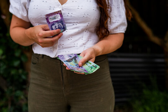 Woman holds New Zealand banknotes while standing in a garden area with trees