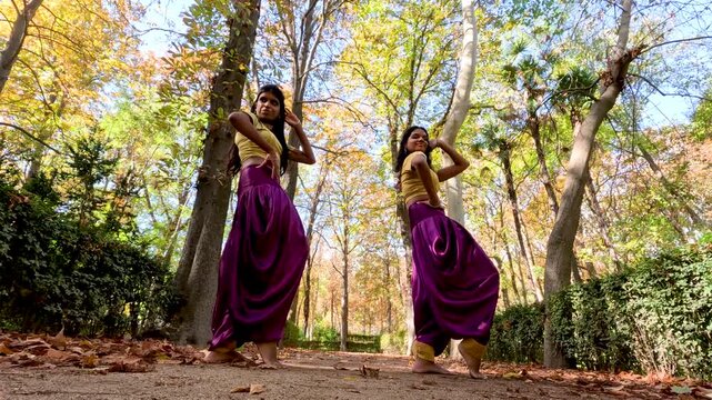 Two young barefoot women in traditional purple and yellow attire performing a synchronized bollywood dance in a beautiful autumn park