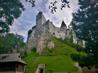 Bran Castle in Romania is perched atop a rocky hillside, partially obscured by tall, leafy trees. The castle features a mix of architectural elements with white stone walls, pointed towers © Julija