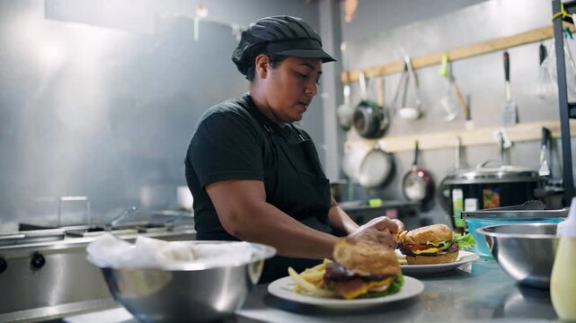 Diligent latina cook working in a professional kitchen, carefully preparing hamburgers with french fries and placing them on plates