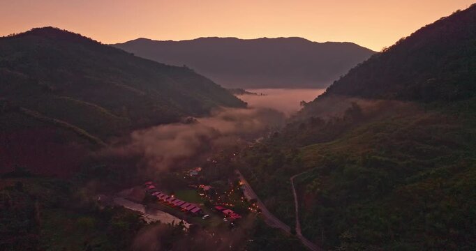 A serene aerial view of a mountain valley at sunrise, with soft mist drifting through the hills and warm lights glowing from a small village below. The scene captures the peaceful atmosphere