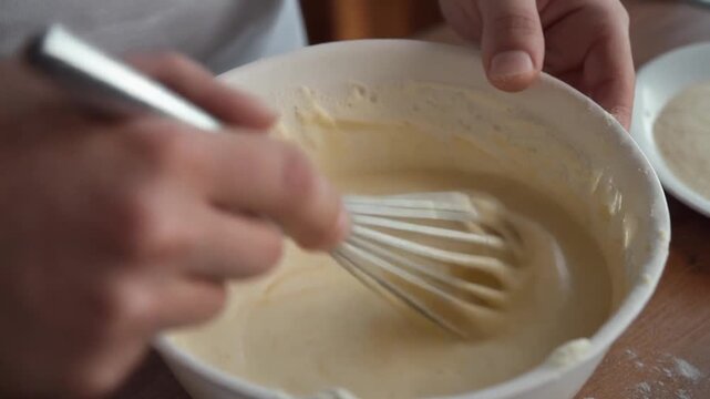 Hands whisking mixture in bowl on table with utensil and plate