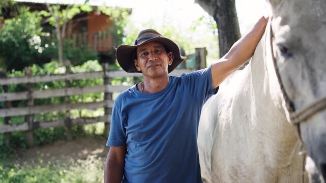 Smiling hispanic farmer wearing a hat standing next to his beautiful white horse. Portrait of an experienced equestrian stableman outdoors