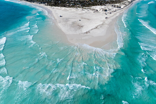 High aerial view of turquoise waves washing over a sharp white sand point into a deep blue ocean