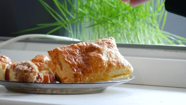 Woman picking up fresh golden pastry from plate. Hand reaching for baked bread roll on kitchen table. Homemade snack being served during breakfast or meal time.