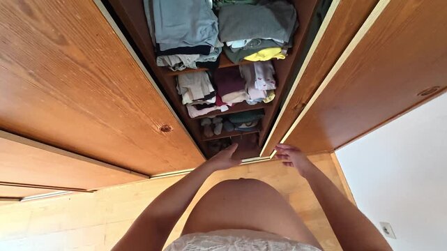 POV of a pregnant woman's hands and body opening a wooden wardrobe with children's clothes inside, looking up and down and taking children's coats in her arms