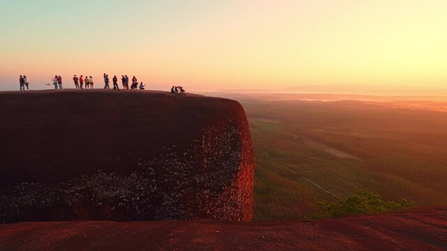 A breathtaking dawn scene featuring small groups of tourists standing atop a giant cliff, backlit by warm sunlight and surrounded by panoramic mountain and forest views.