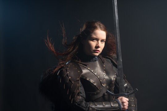 A fierce warrior woman with fiery hair and a sword stands ready against a dark background, clad in detailed armor.