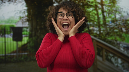 Woman with glasses and curly hair wearing red sweater holds hair and opens mouth wide in forest; surprise amazement.
