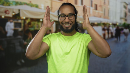 Naklejka premium Man with hands framing face on a restaurant terrace smiling in a bright green tshirt and glasses, casual outdoor scene;joy playfulness.