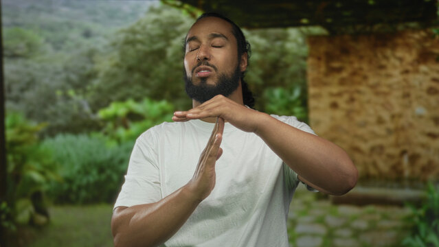 Man hands making time out gesture in forest near stone building and green field, wearing white t shirt; calm pause.