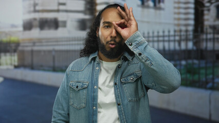 Fototapeta premium Man makes ok sign to eye on street near building fence wearing denim jacket and white t shirt; playful curiosity.
