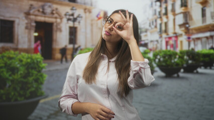 Fototapeta premium Woman with glasses makes ok sign by eye on cobblestone street in front of historic building and planters; confidence.