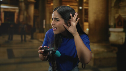 Woman holds gamepad and cups ear with hand while squinting and listening inside a building  concentration challenge determination. © Krakenimages.com