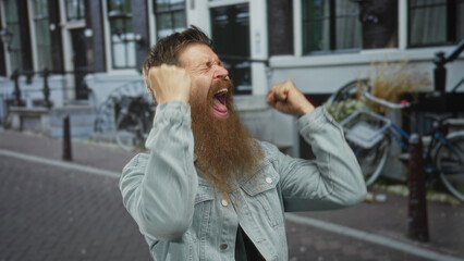 Fototapeta premium Bearded man in denim jacket clenches fists on a busy city street with brick buildings behind him; excitement.