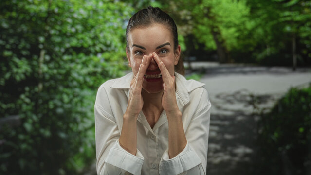 Young hispanic woman wearing white shirt cups her hands around her mouth shouting on sunlit street; public warning.
