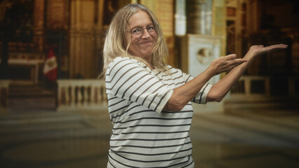 Fototapeta premium Senior woman presenting open palms and smiling in a striped shirt inside an ornate building; grace contentment.