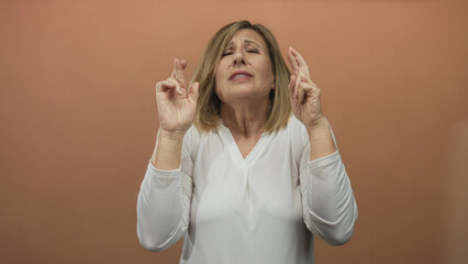 Middle aged woman praying with clasped hands and closed eyes in a peach studio with tense...