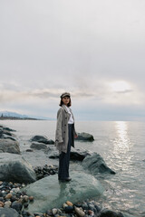 Fototapeta premium Young woman in casual outfit stands on rocky shore near calm sea under cloudy sky. Concept of solitude, nature, and peaceful outdoor lifestyle with soft natural light.