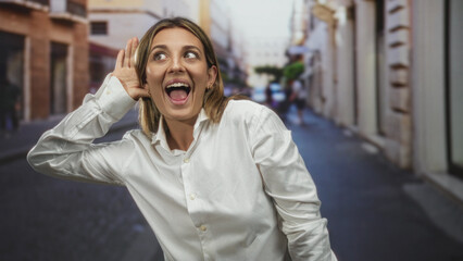 Obraz premium Woman with hand cupped to ear and mouth open on a city street wearing a white shirt, leaning forward as if listening intently; surprise curiosity.