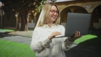 Woman with red glasses wearing white shirt holding laptop waves hand to camera in building courtyard; joy connection.
