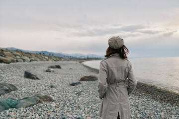 Woman in grey coat and hat stands on rocky beach looking at calm sea under cloudy sky. Concept for solitude, nature, autumn, and peaceful outdoor moment. © SHOTPRIME STUDIO